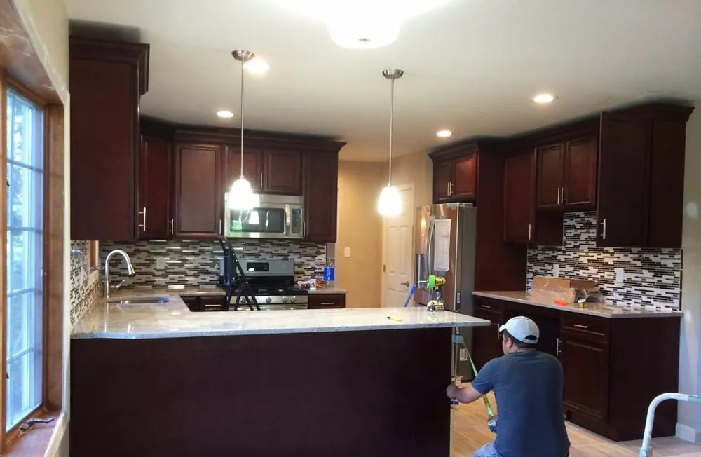 Kitchen with cherry cabinets, glass mosaic backsplash and granite peninsula while a worker finishes installation.