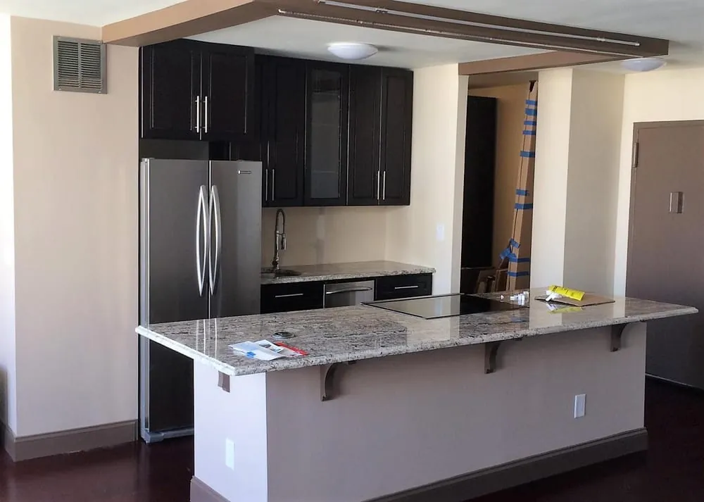 Condo kitchen with espresso cabinets, stainless fridge, granite peninsula with corbels and cooktop, packing materials nearby.