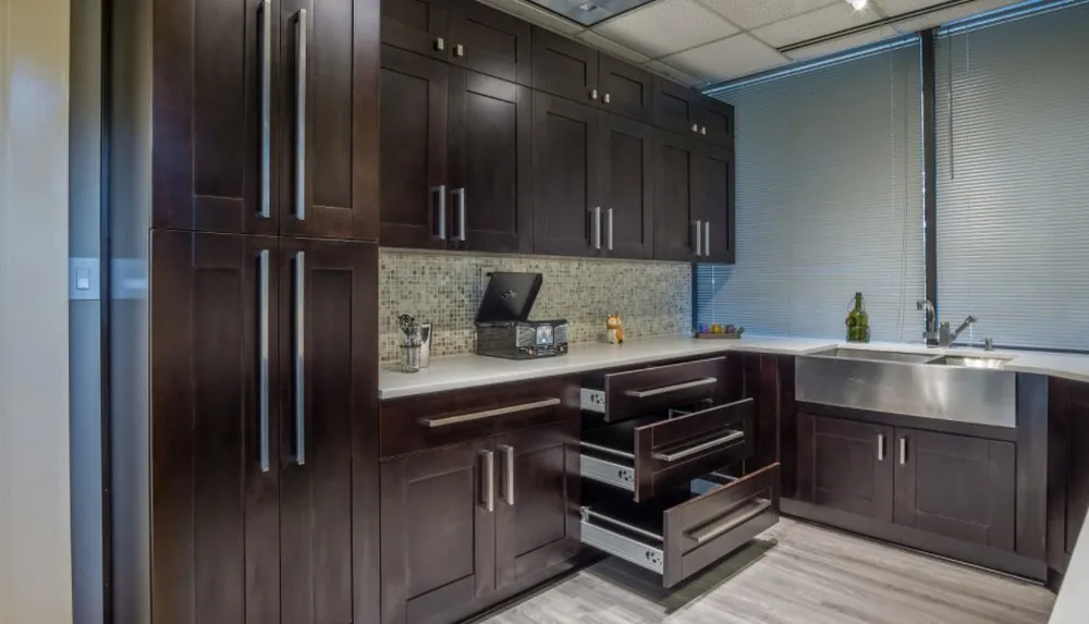 Espresso shaker cabinets with open drawers, white counters, glass mosaic backsplash and a stainless apron sink by blinds.