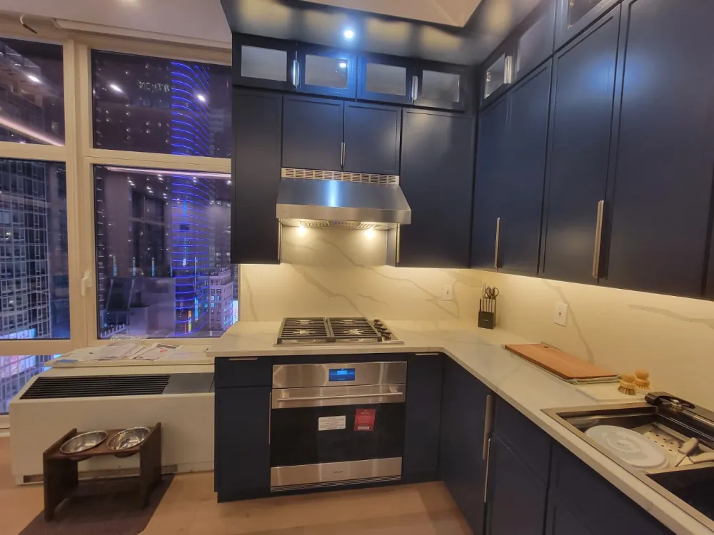 Navy shaker cabinets with glass-front uppers, stainless hood over gas cooktop, wall oven and white quartz backsplash.