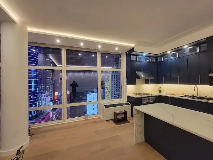 Apartment kitchen with navy cabinets, marble-look waterfall island, white oak floor and floor-to-ceiling windows at night.