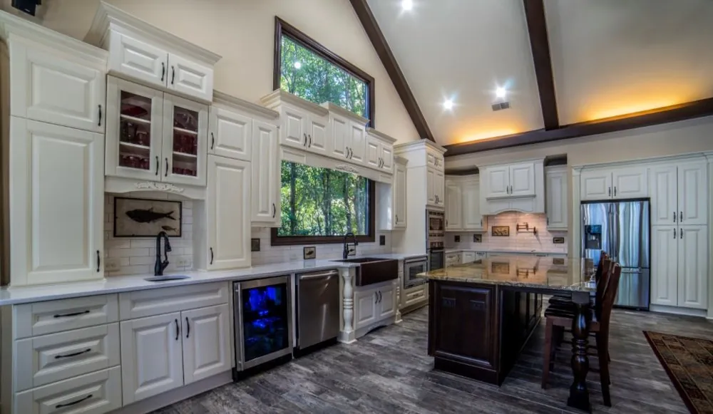 Vaulted kitchen with cream raised-panel cabinets, copper farmhouse sink, large window, dark wood island and gray plank floor.
