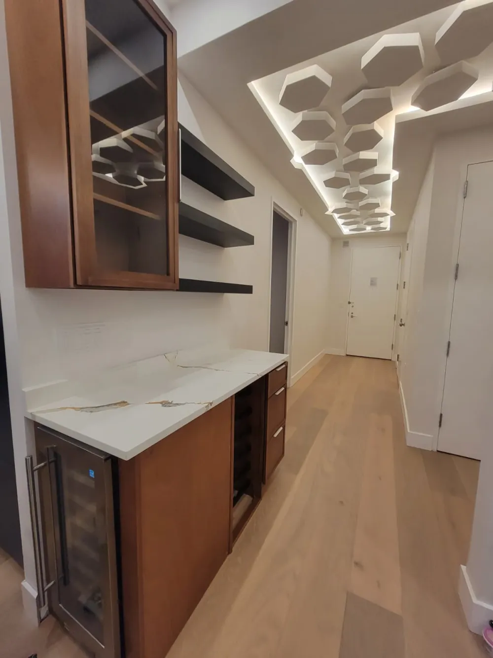 Wet bar with wood cabinets, wine fridge, wine rack, gold-veined quartz top and floating shelves along a hallway.