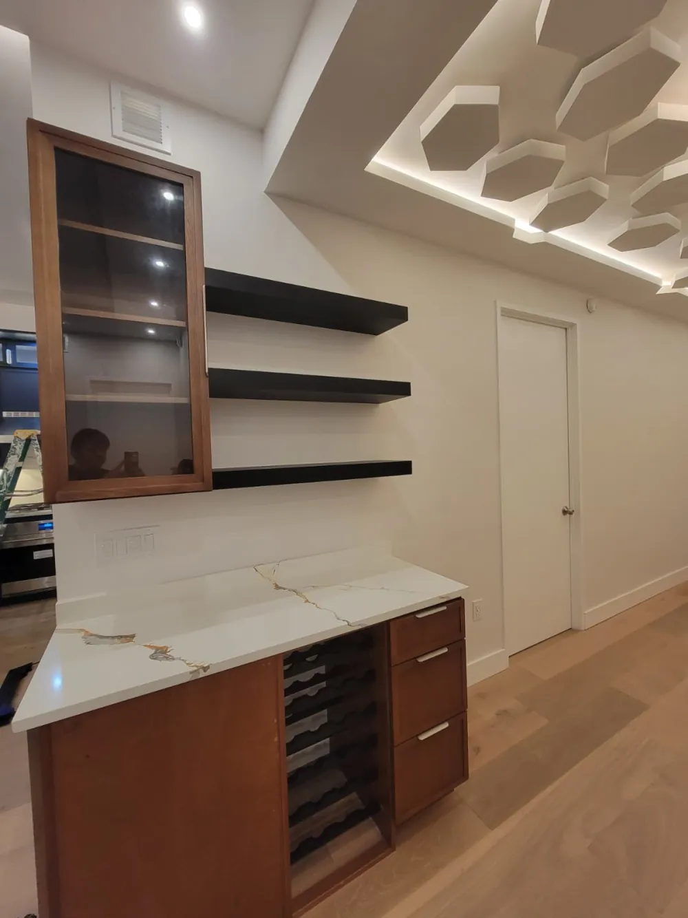 Wet bar with wood cabinets, built-in wine rack, gold-veined white quartz top, glass upper cabinet and black floating shelves.
