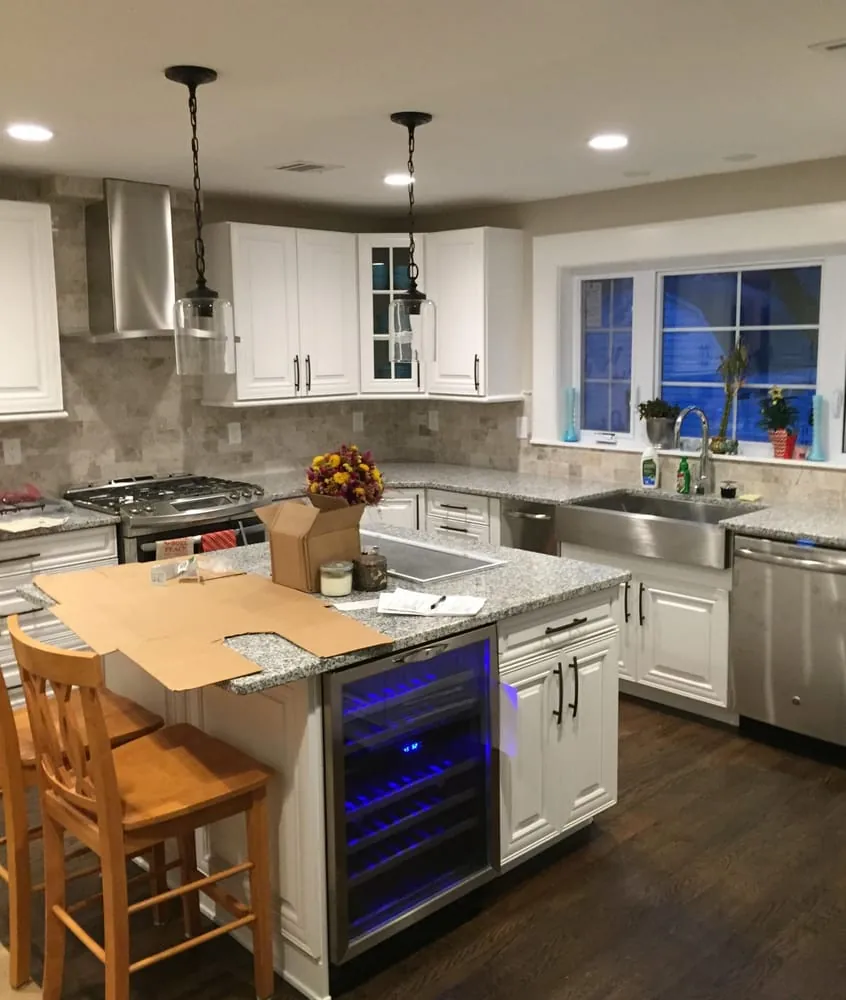 White kitchen with gray granite island holding a wine fridge, farmhouse sink under windows, chimney hood and glass pendants.