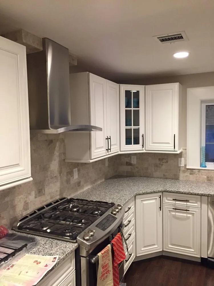 White raised-panel corner cabinets with a glass door, gray granite counters, travertine backsplash, chimney hood, gas range.