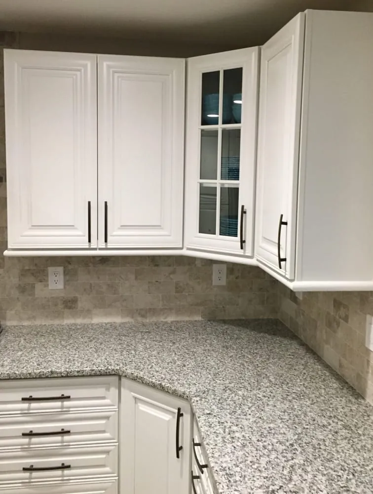 White raised-panel upper cabinets with a glass-front corner cabinet over gray granite counters and travertine backsplash.