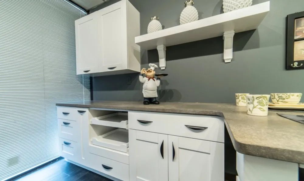White shaker cabinets with roll-out trays, gray laminate counter, open white shelf with brackets and a dark gray wall.