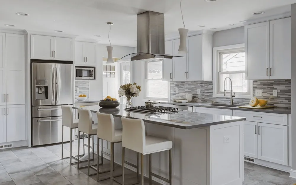 White shaker kitchen with island cooktop under a stainless chimney hood, gray quartz tops, glass mosaic backsplash, stools.