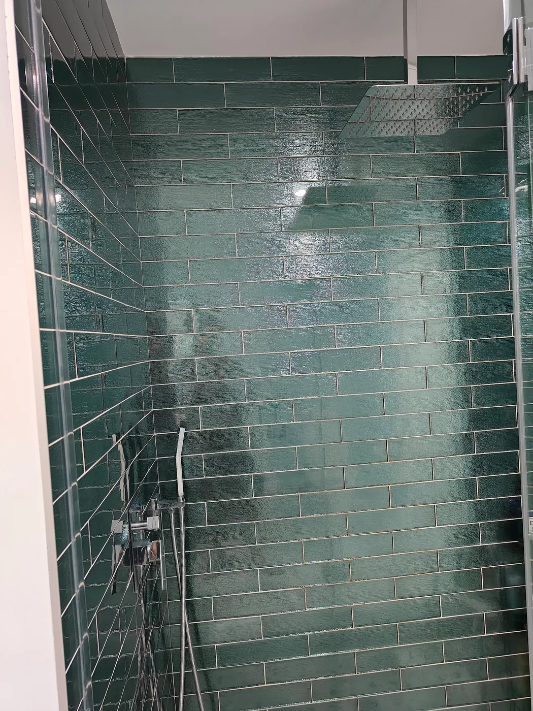 Shower lined with glossy green subway tile, with a ceiling rain shower head, handheld sprayer and glass enclosure